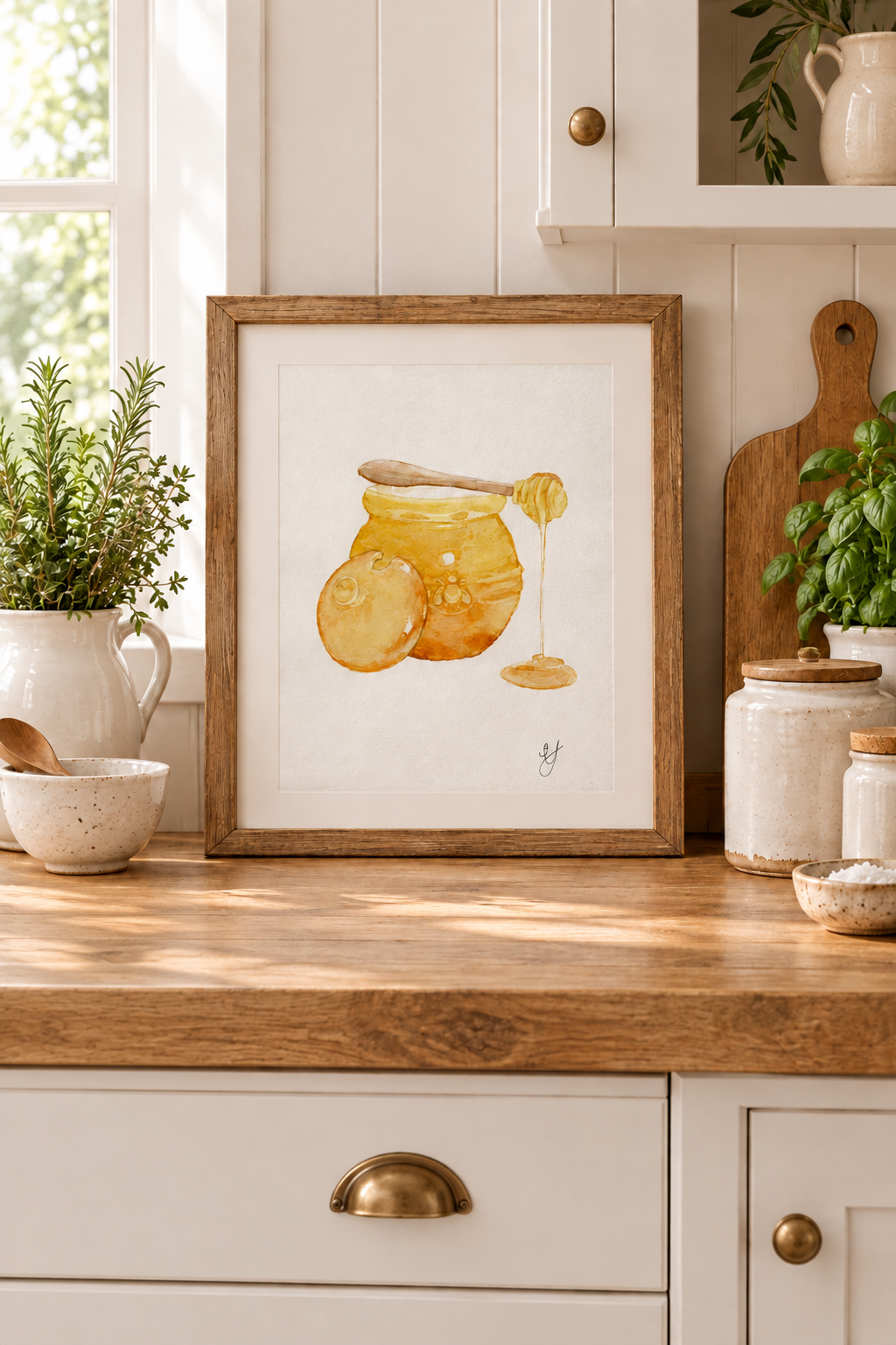 A framed watercolor art print of honey jars and a honey dipper, styled on a rustic wooden kitchen counter with white cabinets, ceramic jars, and fresh greenery.